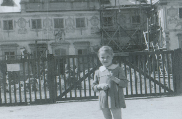 Dziecko stoi z kwiatkiem w ręku. W tle fragment pałacu. Pałac jest w remoncie, przy fasadzie postawiono rusztowania. 