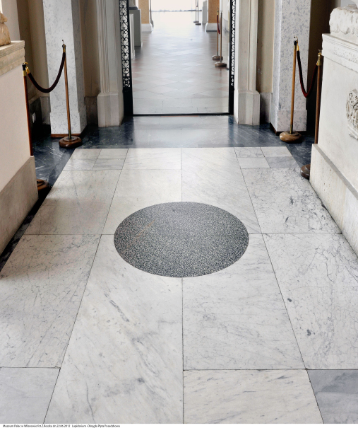 Zdjęcie przedstawia korytarz w pałacu. Na pierwszym planie posadzka z okrągłym, szarym elementem kamiennym. Jest to najcenniejszy fragment posadzki w Lapidarium.