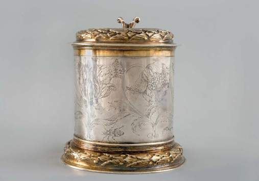 Srebrny kufel ze złotymi motywami zdobiony grawerunkiem. Na jego brzuścu znajdują się kwiaty oraz owady. 

