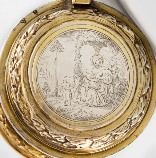 Okrągła, złocona pokrywa od kufla. Wieko grawerowane przedstawia siedząca kobietę, która karmi dziecko piersią. Drugie małe dziecko stoi przed nią i czyta książkę. Scena rozgrywa się na tle pejzażu. Jest to alegoria wieku dziecięcego. 