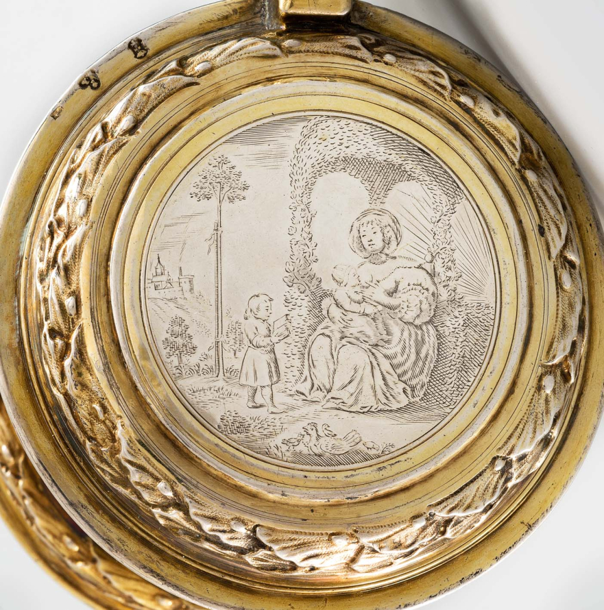 Okrągła, złocona pokrywa od kufla. Wieko grawerowane przedstawia siedząca kobietę, która karmi dziecko piersią. Drugie małe dziecko stoi przed nią i czyta książkę. Scena rozgrywa się na tle pejzażu. Jest to alegoria wieku dziecięcego. 