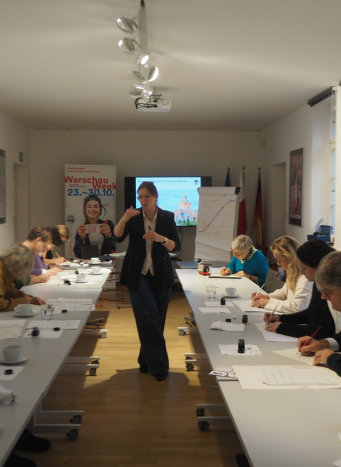 Młoda kobieta stojąca w centrum wyjaśnia zagadnienie z kaligrafii grupie osób usadzonych przy stołach po dwóch stronach.