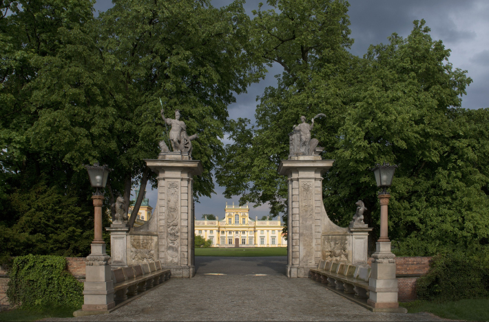 Zdjęcie przedstawia bramę główną Pałacu Wilanowskiego i drzewa. W tle widoczna fasada pałacu.