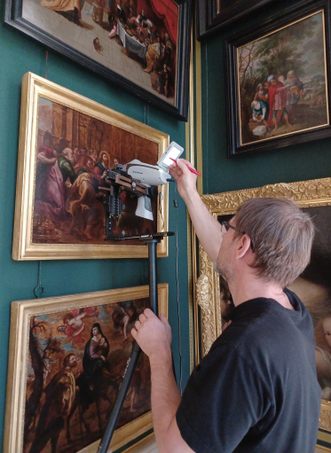 Mężczyzna ze specjalistycznym sprzętem bada obrazy w złotych ramach powieszonych na ścianie.